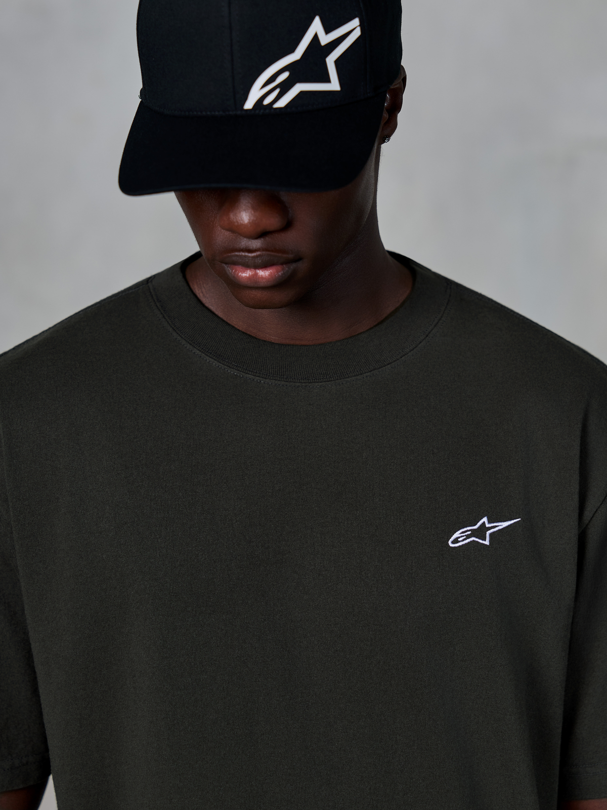 Alpinestars Essentials Ageless SS Tee, Kurzarm-T-Shirt, Off Black, mit kleinem weißen Alpinestars-Logo auf der Brust, getragen mit einer schwarzen Baseballkappe mit Logo, lässiger Motorsport-Streetwear-Stil