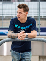 Alpinestars Collared CSF Tee, Kurzarm-T-Shirt, Marineblau, petrolfarbener Alpinestars-Schriftzug und weißes Stern-Logo, getragen von einem Mann, der an einem Metallgeländer an einer Rennstrecke lehnt, lässige Motorsport-Streetwear