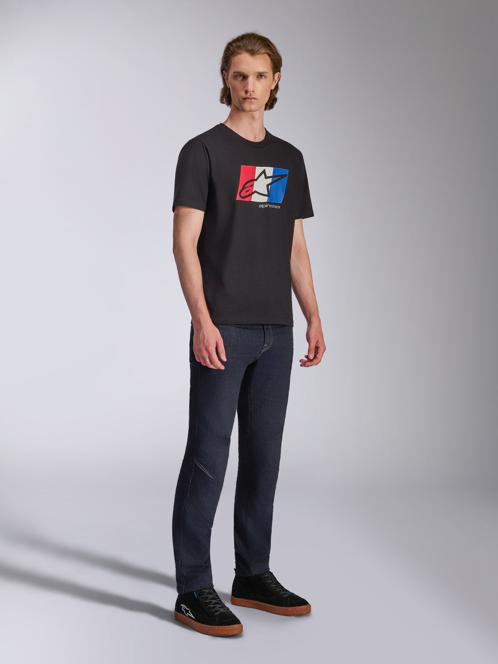 Alpinestars Global CSF Tee, Kurzarm-T-Shirt, Schwarz mit rot-weiß-blauer Logo-Grafik, lässige Motorsport-Streetwear, Ganzkörperaufnahme eines Models in dunkler Jeans und schwarzen Sneakern