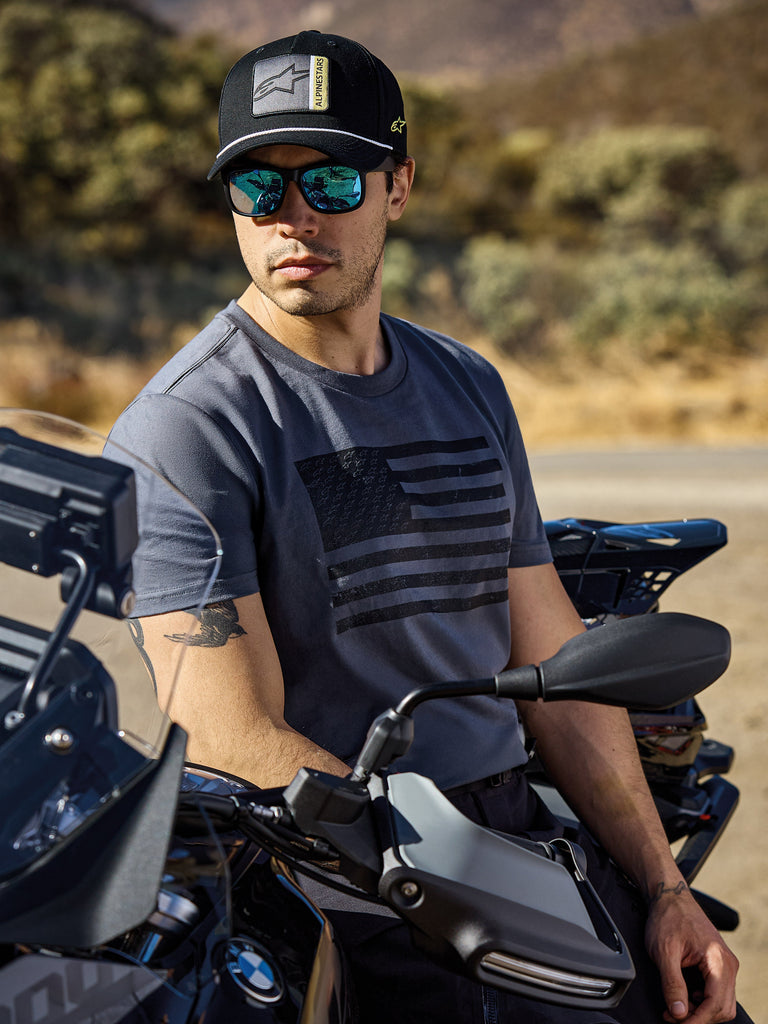 Alpinestars Waving CSF Tee, Kurzarm-T-Shirt, Charcoal, mit einer schwarzen Grafik einer wehenden Flagge auf der Brust, getragen von einem Mann auf einem Motorrad in einer sonnigen Außenumgebung, lässiger Motorsport-Lifestyle