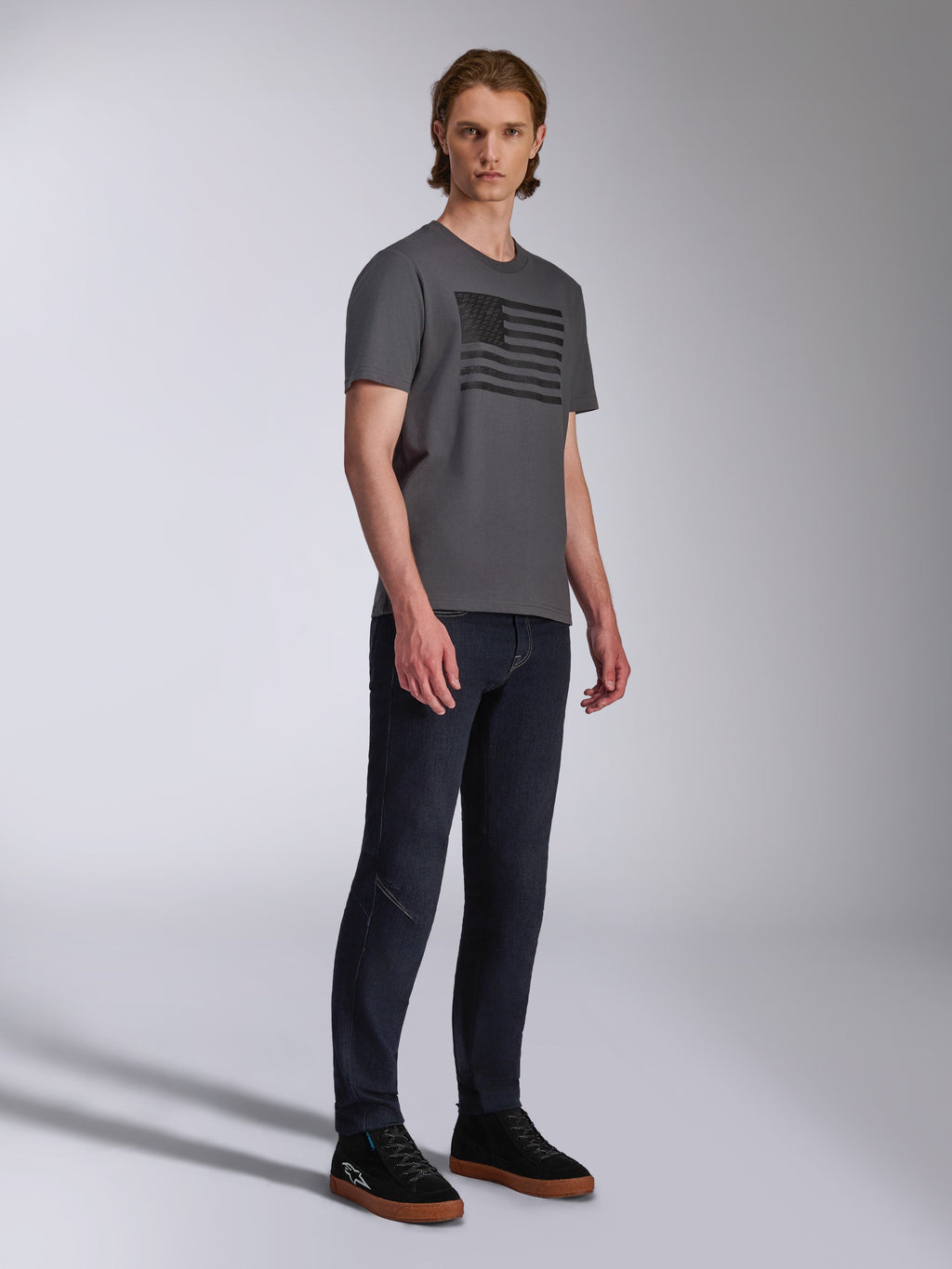 Alpinestars Waving CSF Tee, Kurzarm-T-Shirt, Charcoal, Grafik mit schwarzer wehender Flagge, getragen von einem Model mit dunkler Jeans und Sneakern, Casual Motorsport Streetwear
