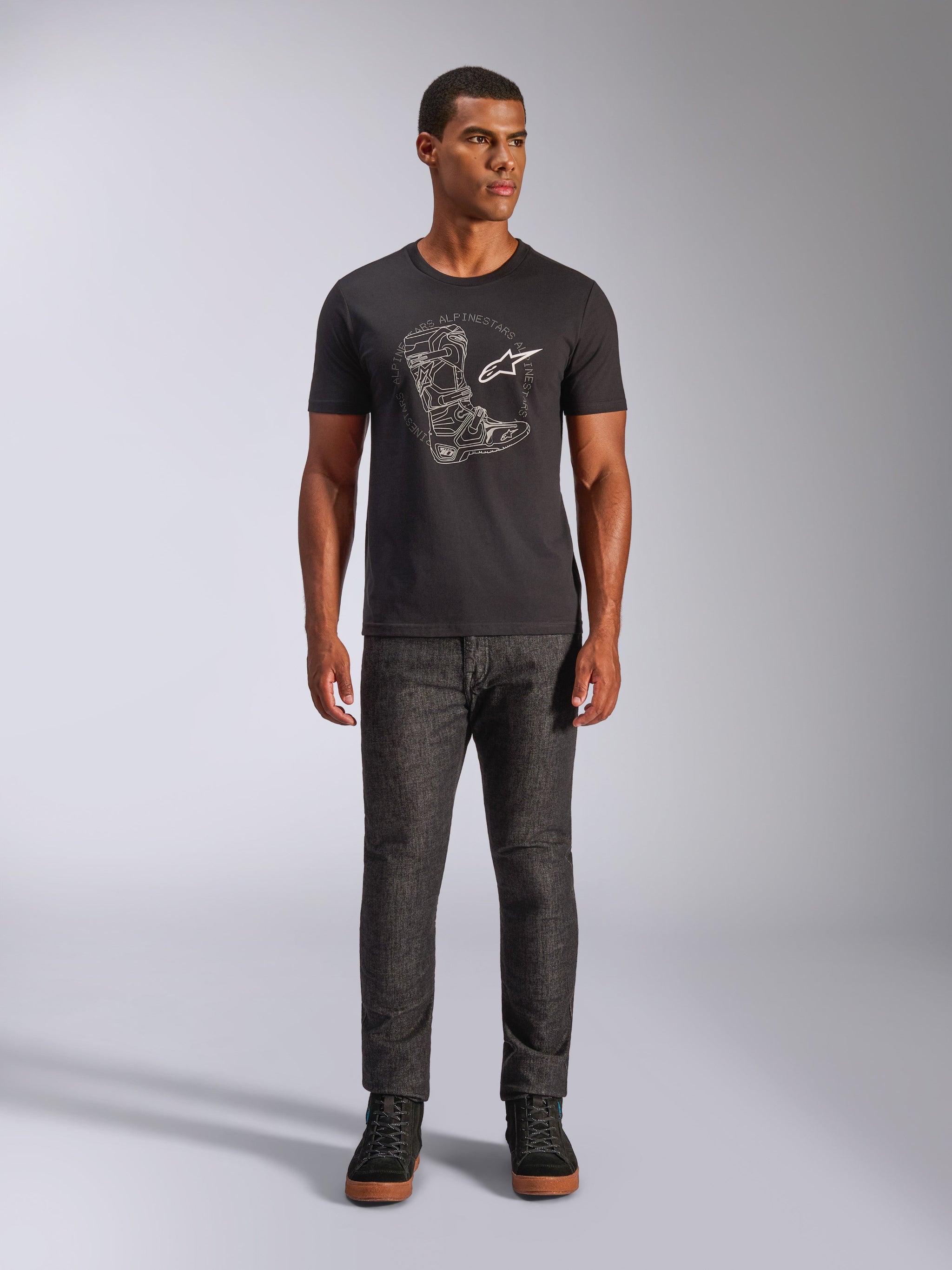 Alpinestars Tech Boot CSF Tee, Kurzarm-T-Shirt, Schwarz, mit weißer Grafik eines Motocross-Stiefels und Logo auf der Brust, getragen von einem Model mit dunkelgrauer Hose und schwarzen Sneakern, lässiges, vom Motorsport inspiriertes Design