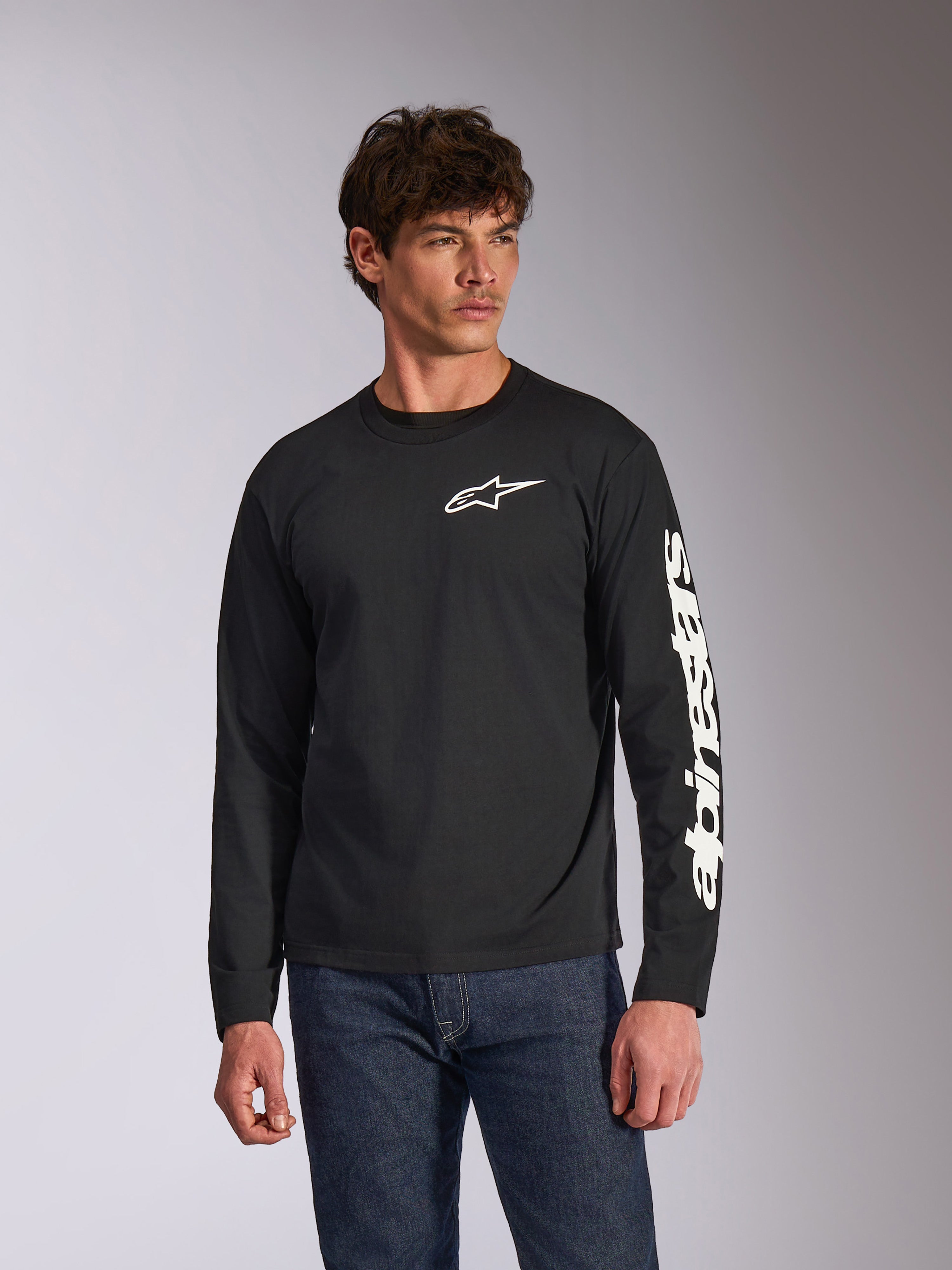 Alpinestars Trackside CSF Langarmshirt, Schwarz, weißes Alpinestars-Logo auf der Brust und Branding am Ärmel, getragen von einem Model mit blauen Jeans, lässige Motorsport-Streetwear
