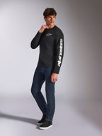 Alpinestars Trackside CSF Tee, Langarmshirt, Schwarz, weißes vertikales Alpinestars-Branding auf dem Ärmel und Brust-Logo, getragen von einem männlichen Model mit dunkelblauen Jeans und Sneakern, lässige Motorsport-Streetwear