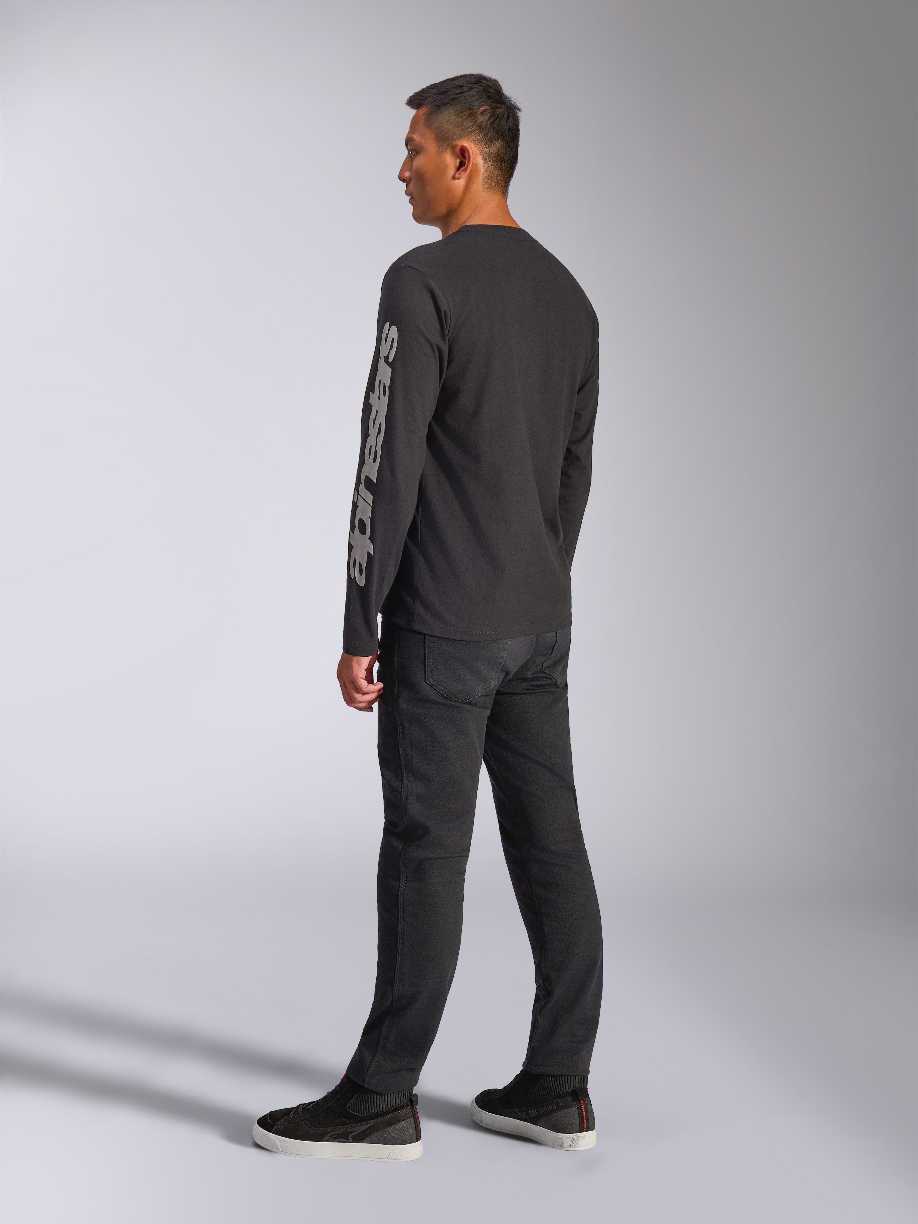 Alpinestars Trackside CSF Langarm-T-Shirt, Schwarz, mit großem grauem Alpinestars-Logo auf dem linken Ärmel, getragen von einem Modell mit schwarzer Hose und lässigen Sneakern, Motorsport-inspirierte Streetwear.