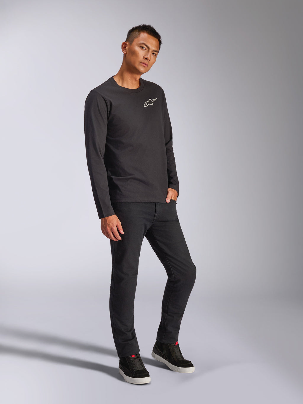 Alpinestars Trackside CSF Tee, Langarm-T-Shirt, Schwarz, weißes Astars-Logo auf der Brust, getragen von einem Model mit schwarzer Hose und Sneakern, lässige, vom Motorsport inspirierte Streetwear