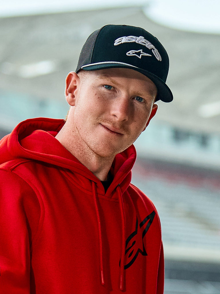 Mann in einem roten Alpinestars-Hoodie und der Agree Trucker Mütze in Schwarz, mit weiß gestickten Astars-Logos, einem weißen Kordel-Detail am Schirm und einer atmungsaktiven Mesh-Rückseite, lässiges Lifestyle-Setting