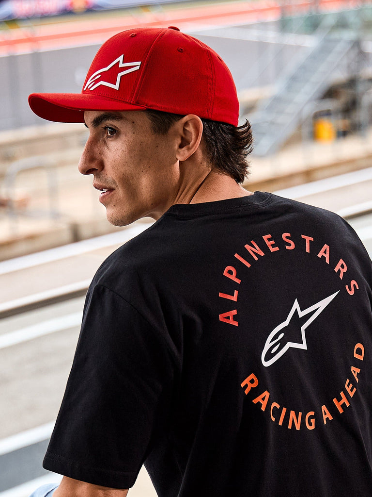 Alpinestars Corp Shift Multi Hat, Red curved bill cap with white 3D Astars logo, worn with a black Alpinestars t-shirt featuring 'Racing Ahead' graphic, lifestyle motorsport setting at a racing circuit