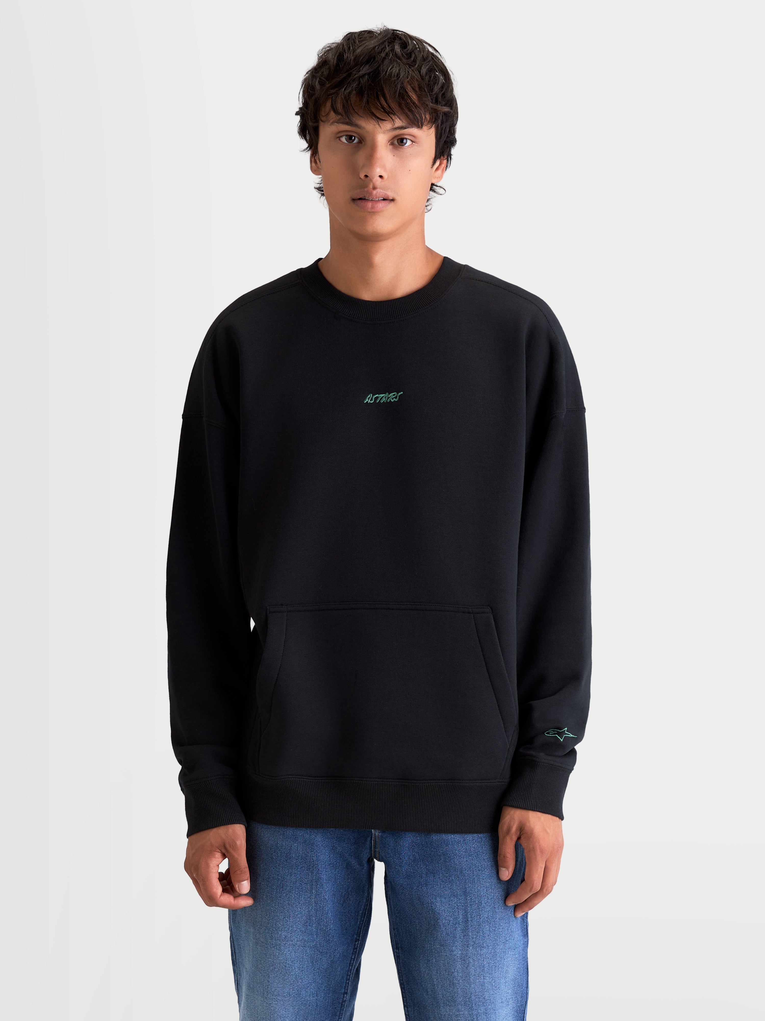Alpinestars Guise Oversized Crew, schwarzer Sportswear-Pullover an einem Modell, Unisex, Oversized-Passform mit Kängurutasche, blaugrün gestickte Astars-Logos auf Brust und Ärmel, Fleece aus Baumwolle und recyceltem Polyester.