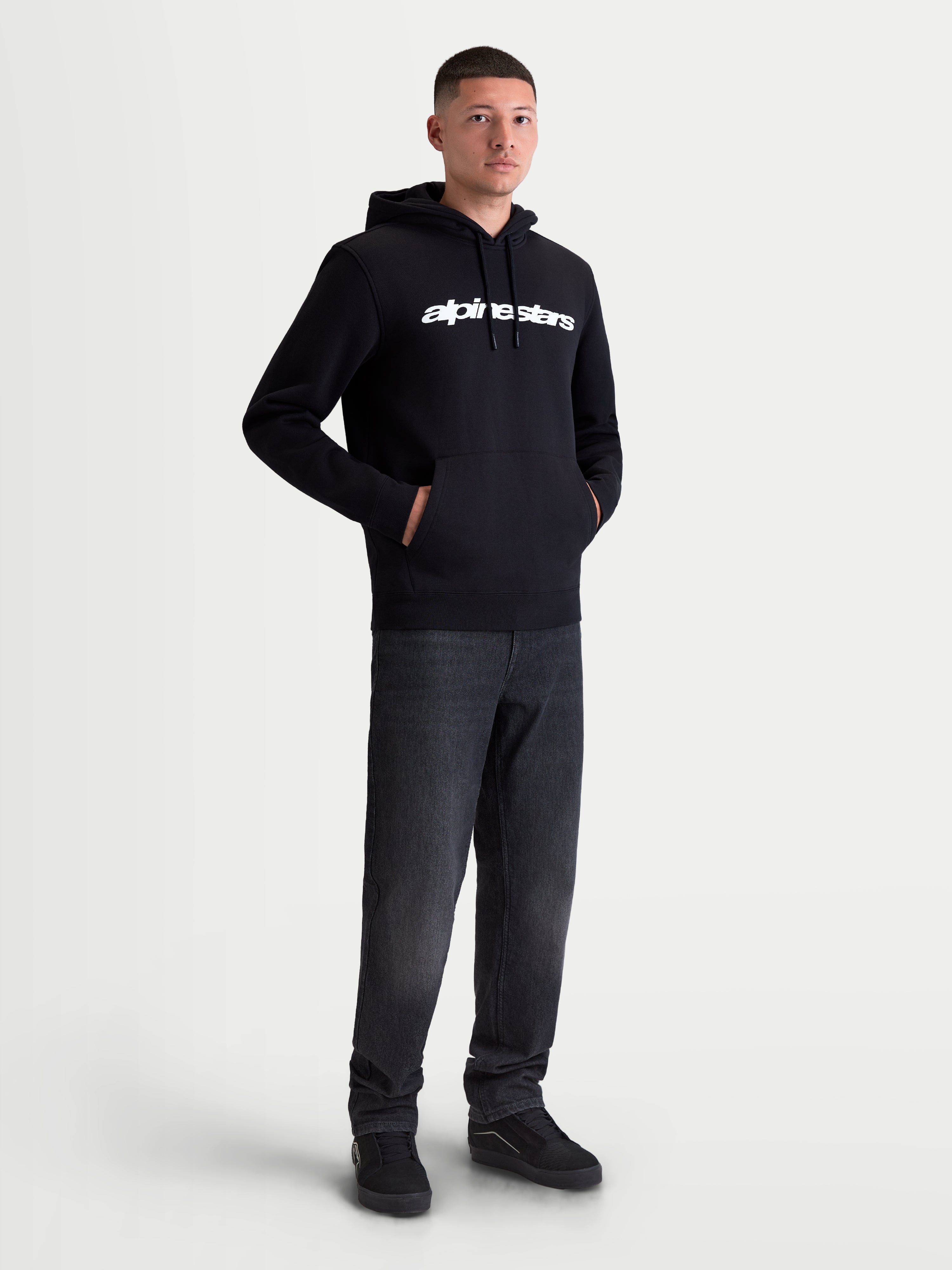 Alpinestars Race Wordmark Hoodie, Herren Kapuzenpullover aus Fleece, Schwarz und Weiß, mit markantem weißem Brustlogo, Kapuze mit Kordelzug und Kängurutasche vorne. Hergestellt aus einer Baumwollmischung mit recyceltem Polyester für den Alltag, am Modell präsentiert.