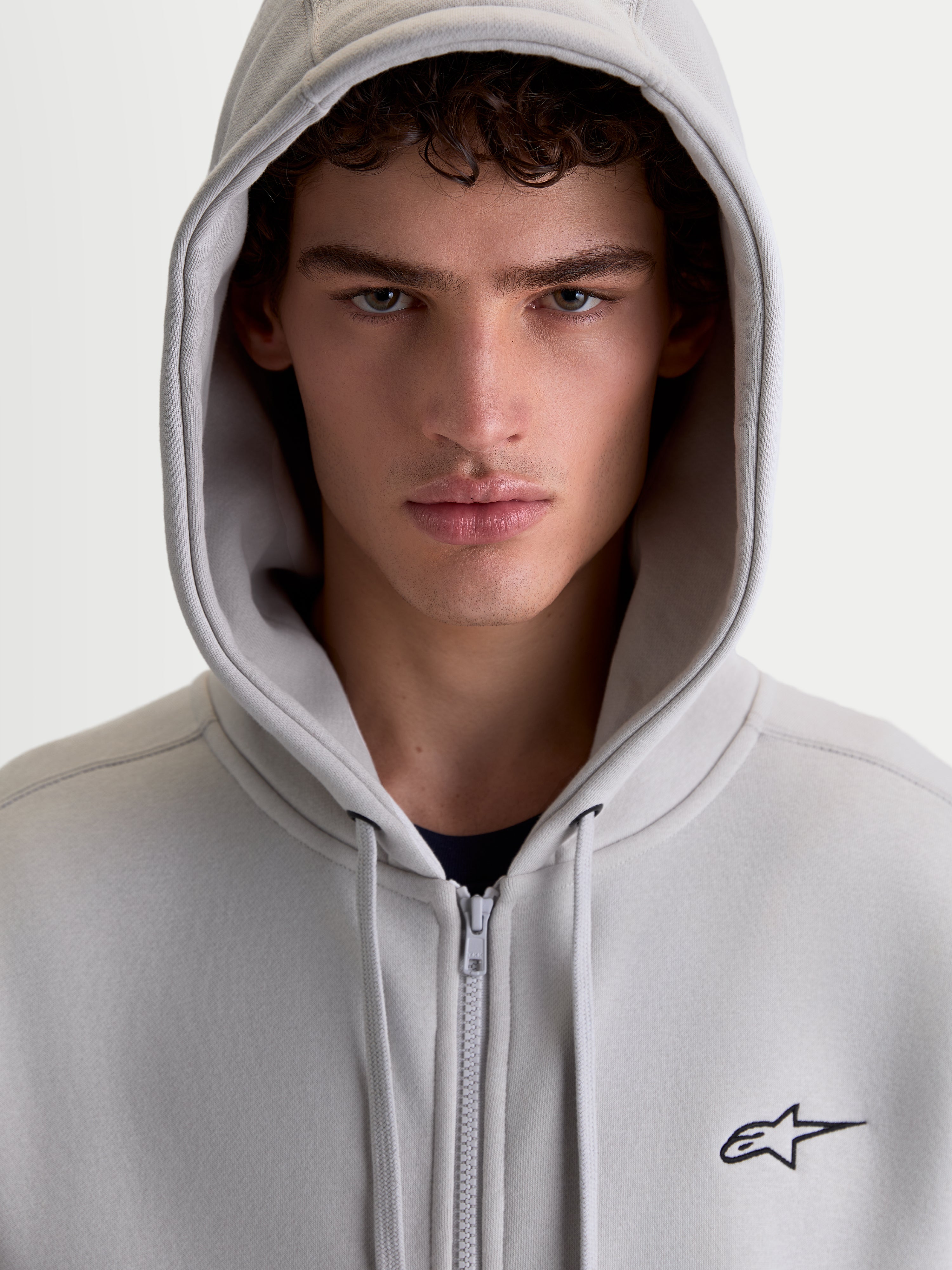 Alpinestars Entity Oversized Hoodie, Sportswear-Zip-Hoodie, Zementgrau, mit Vislon-Frontreißverschluss und gesticktem Astars-Logo, Nahaufnahme-Porträt eines Models mit aufgesetzter Kapuze.