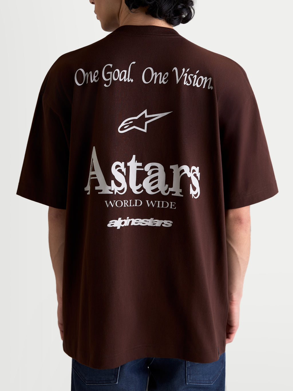 Alpinestars Post Oversized CSF T-Shirt, braunes Kurzarm-Sportswear-T-Shirt, Rückenansicht am Model mit weißen 'One Goal. One Vision.' und stilisierten 'Astars World Wide' Grafiken, lockere Oversized-Passform aus Baumwoll-Polyester-Mischgewebe.