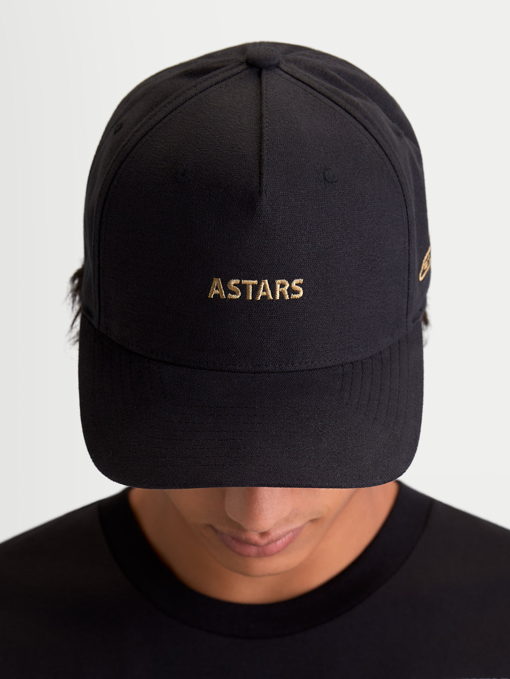 Alpinestars County Canvas Kappe, schwarze Sportswear-Kappe mit goldener ASTARS-Logo-Stickerei, gebogenem Schirm und strukturierter Krone, Unisex Motorsport-inspirierte Kappe aus Oxford-Peach-Gewebe, Frontansicht auf Modell.