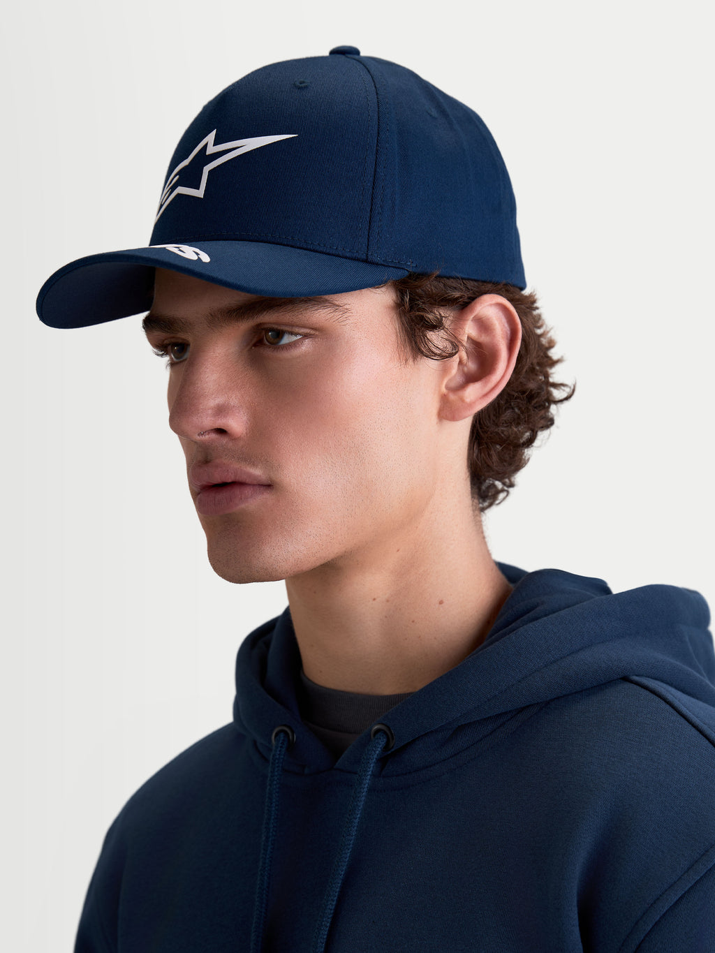 Alpinestars Arclight Kappe, dunkelblaue Sportswear-Kappe für Herren und Damen, mit weißem Astars Puff-Print-Logo auf der strukturierten Krone und gebogenem Schirm. Seitenprofil-Ansicht am Modell, klassische Motorsport-Passform aus strapazierfähigem Baumwoll-Twill.
