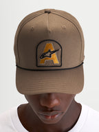 Alpinestars Acme Snapback Kappe in Walnussbraun mit gebogenem Schirm, vorne aufgesticktem Astars-Logo-Patch und schwarzem Kordelbesatz, getragen von einem Model