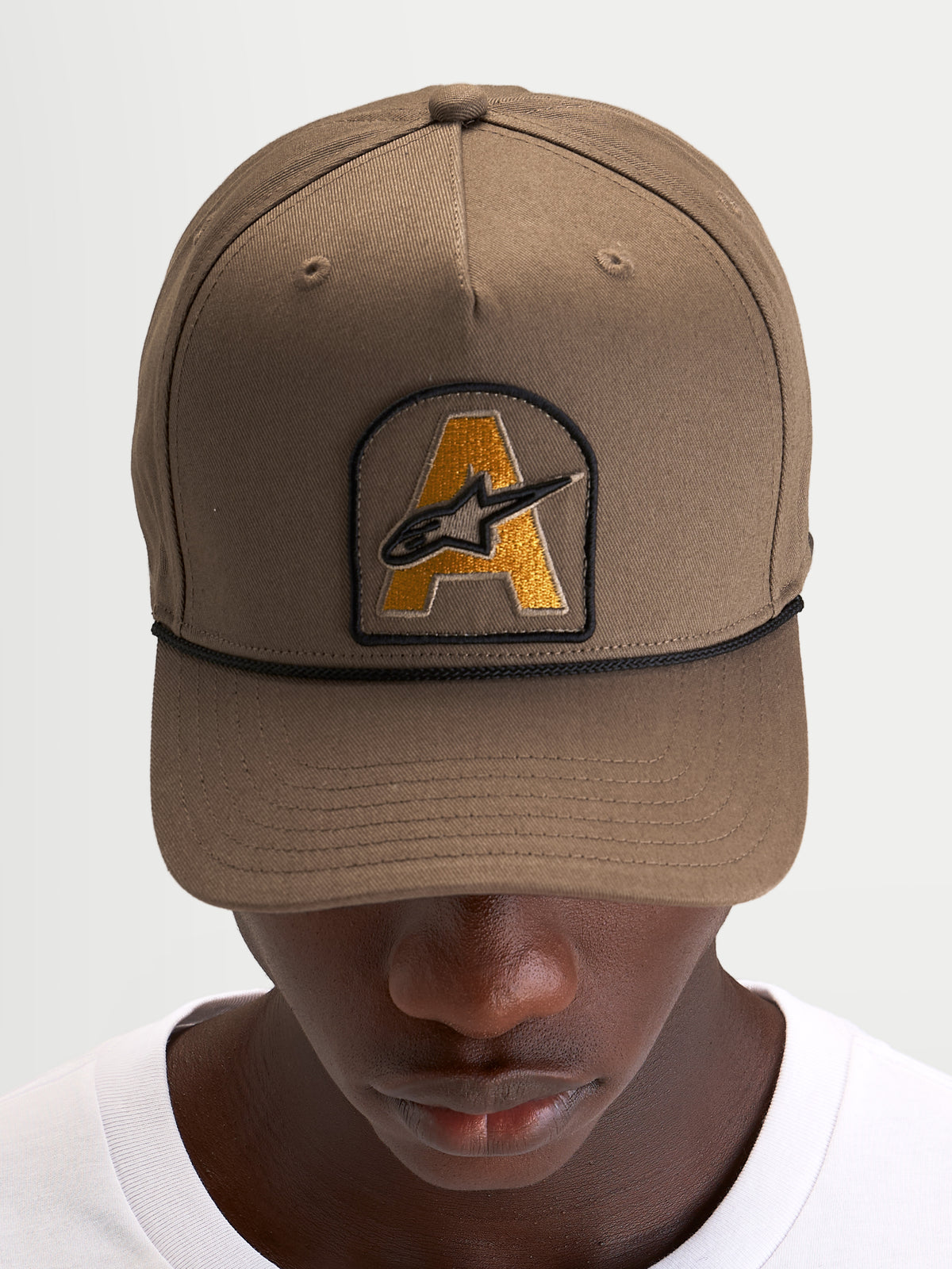 Alpinestars Acme Snapback Kappe in Walnussbraun mit gebogenem Schirm, vorne aufgesticktem Astars-Logo-Patch und schwarzem Kordelbesatz, getragen von einem Model