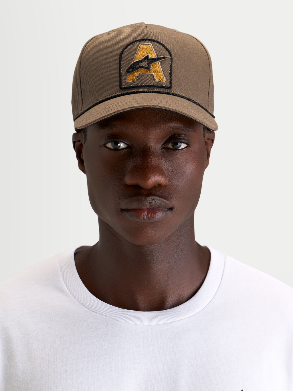 Alpinestars Acme Snapback Kappe, braune Sportswear-Kappe getragen von einem Model, mit strukturierter Krone, gebogenem Schirm und Astars-Logo-Stickpatch vorne mit Seilbesatz