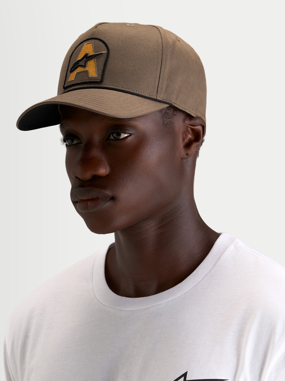 Alpinestars Acme Snapback Kappe, braune Sportswear-Kappe in Walnussbraun mit gebogenem Schirm, strukturierter Krone und Astars-Logo-Stickpatch vorne mit Seilbesatz, getragen von einem Model