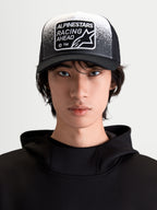 Alpinestars Gravel Trucker-Kappe, Sportswear-Lifestyle-Cap für Herren und Damen, Schwarz und Weiß, Frontansicht am Modell mit gebogenem Schirm, atmungsaktiver Mesh-Rückseite und gewebtem Racing Ahead Patch mit Kieselstruktur-Detail.