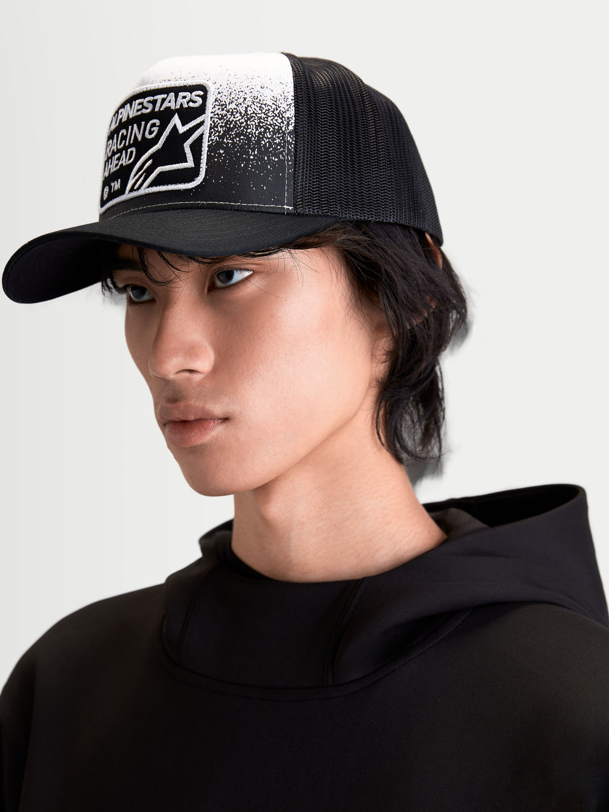 Alpinestars Gravel Trucker-Kappe, Sportswear-Mütze auf Model, Schwarz und Weiß, mit strukturierter Krone, gebogenem Schirm, atmungsaktiver Mesh-Rückseite und gewebtem Patch mit Kieselstruktur-Grafik und Alpinestars Racing Ahead Branding.