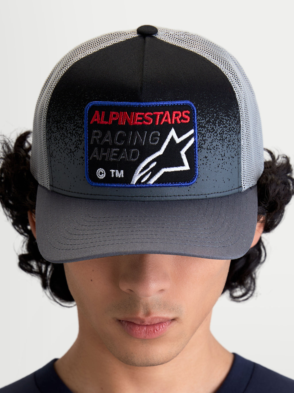 Alpinestars Gravel Trucker-Kappe, Sportswear-Mütze in Dunkelgrau und Schwarz, mit gebogenem Schirm und gewebtem Front-Patch mit Racing Ahead-Text und Astars-Logo, Frontalansicht am Model.