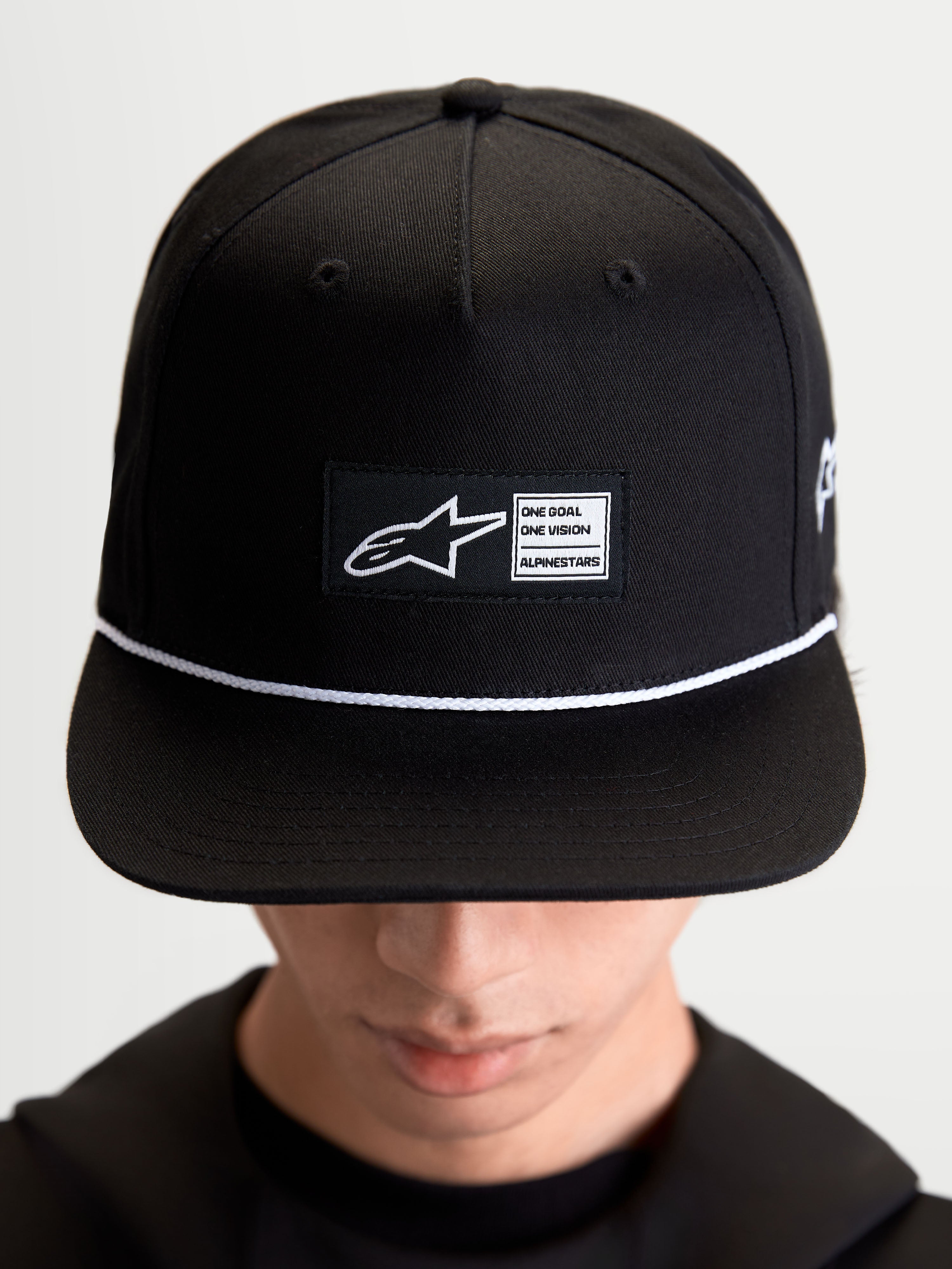 Alpinestars Label Snapback Hat, Sportswear Lifestyle Cap, Unisex, Black and White, Cotton Twill, featuring a structured crown, flat bill, white cord accent, and a front woven label patch with signature Alpinestars branding, shown on-model.