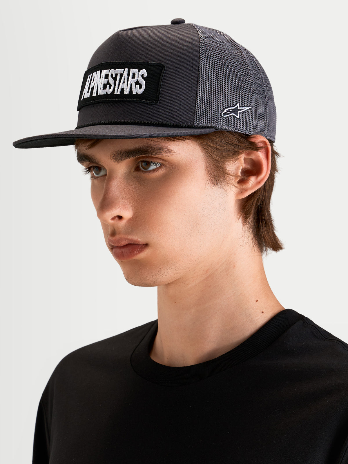 Alpinestars Ten Trucker-Kappe, dunklegraue Trucker-Mütze mit flachem Schirm und gesticktem Alpinestars Wortlogo-Patch vorne sowie Astars-Logo an der Seite, getragen von einem Model
