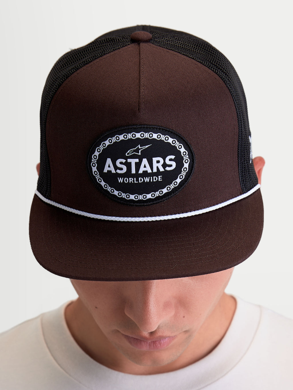 Alpinestars Hotlink Trucker-Kappe, motorsport-inspirierte Sportswear-Kappe, am Modell gezeigt, Braun und Schwarz, mit strukturierter Krone, flachem Schirm mit weißem Kordel-Detail und Astars Worldwide Logo-Patch mit Fahrradketten-Rand.