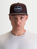 Alpinestars Hotlink Trucker-Kappe, Motorsport-inspirierte Sportswear-Cap in Braun und Schwarz, mit strukturierter Krone, flachem Schirm mit weißem Kordeldetail und gewebtem Astars Worldwide Logo-Patch, am Modell gezeigt.