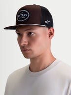 Alpinestars Hotlink Trucker-Kappe, Lifestyle-Sportswear-Kappe getragen am Model, Braun und Schwarz, Twill-Hauptmaterial, strukturierte Krone, flacher Schirm und gewebtes Astars-Logo-Patch mit Fahrradkettenrand.