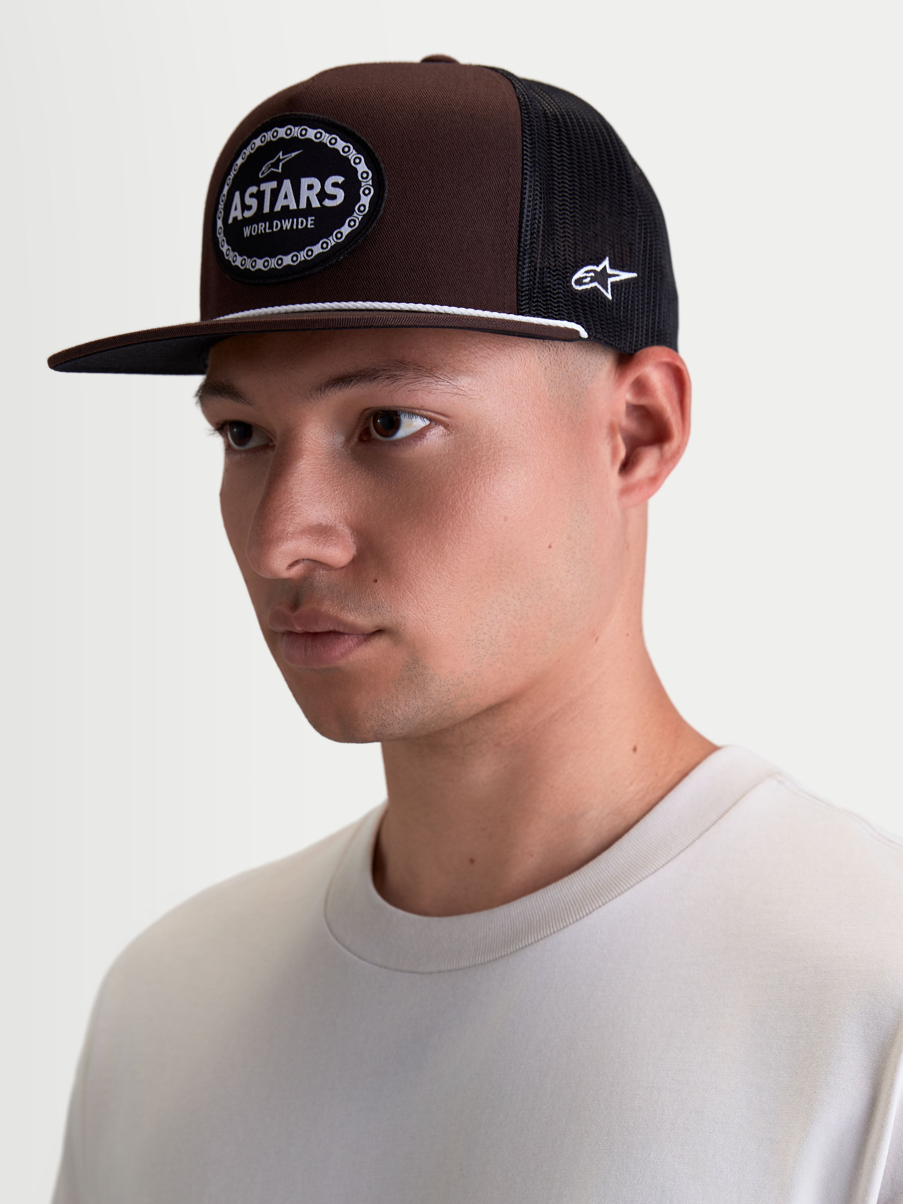 Alpinestars Hotlink Trucker-Kappe, Lifestyle-Sportswear-Kappe getragen am Model, Braun und Schwarz, Twill-Hauptmaterial, strukturierte Krone, flacher Schirm und gewebtes Astars-Logo-Patch mit Fahrradkettenrand.