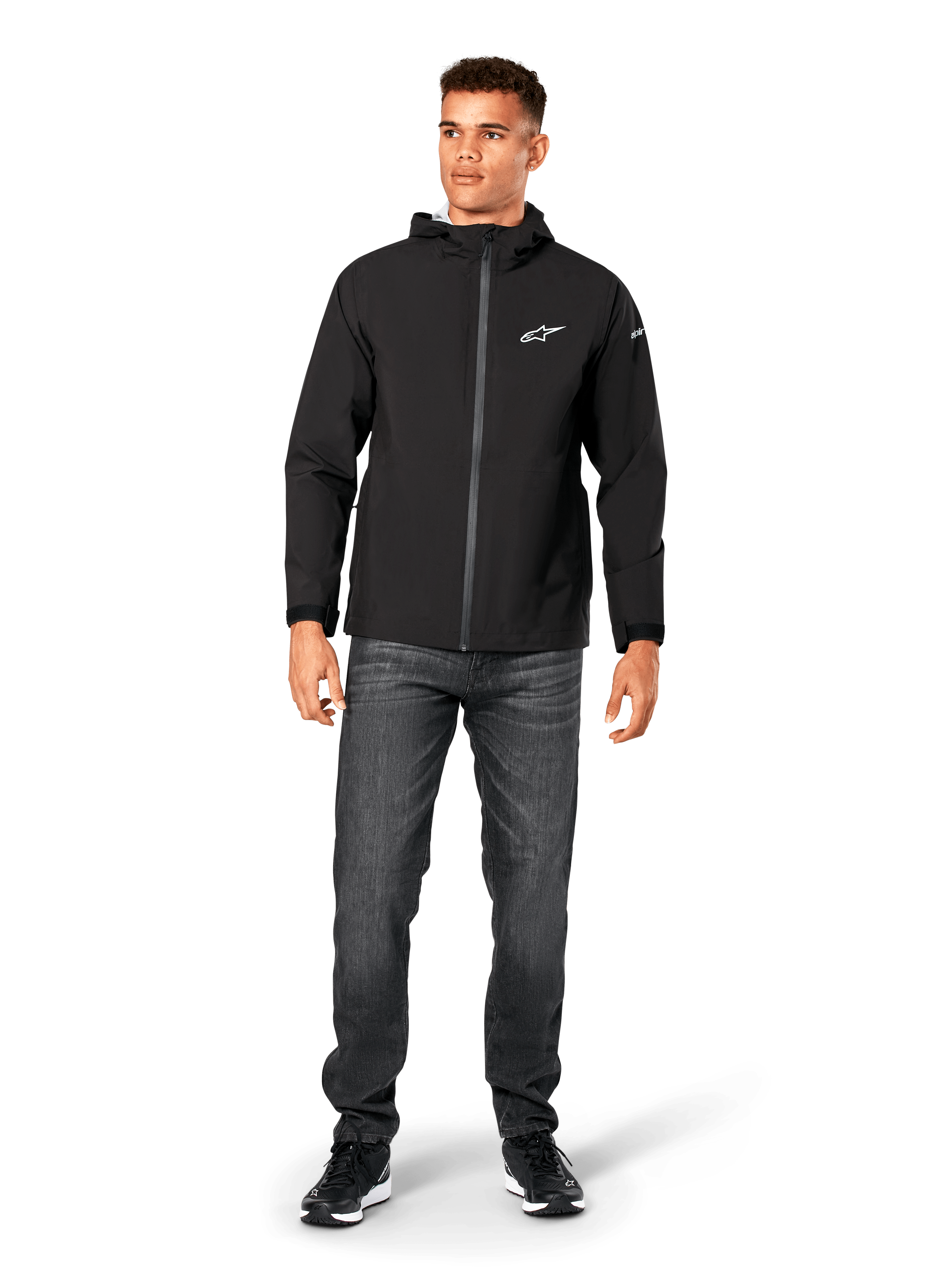 Alpinestars Kascade Regenjacke, Herren-Regenjacke, Schwarz, kombiniert mit dunkelgrauen Jeans und schwarzen Sneakern, mit Kapuzendesign, beschichtetem Frontreißverschluss und weißem Alpinestars-Logo auf der Brust, lässige, vom Motorsport inspirierte Streetwear