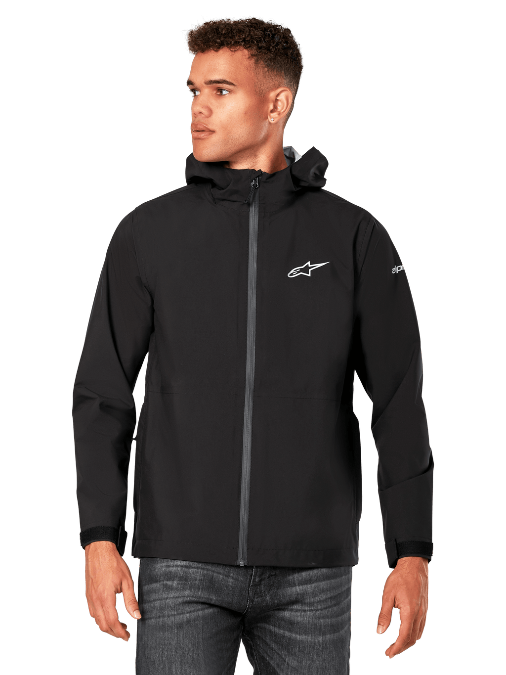 Männliches Model trägt Alpinestars Kascade Regenjacke, Schwarz, weißes Brustlogo und Ärmel-Branding, Regenjacke mit Kapuze, beschichtetem Frontreißverschluss und Klettverschlussbündchen, kombiniert mit dunkler Jeans, lässige Motorsport-inspirierte Streetwear