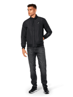 Alpinestars Precedent Jacke, gesteppte Sportjacke, Schwarz, getragen mit dunkelgrauer Jeans und schwarzen Sneakern, mit schrägem Steppmuster, niedrigem Stehkragen, Rippstrickbündchen an Ärmeln und Saum, weißem Logo-Patch auf der Brust und Stickerei am Ärmel.