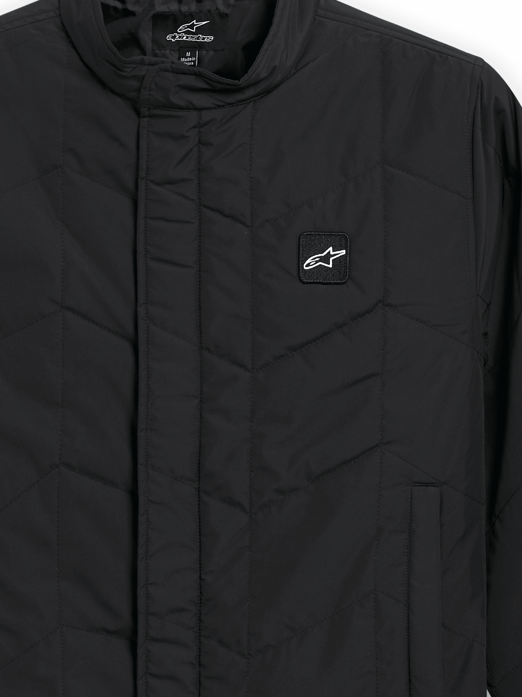 Alpinestars Precedent Jacke, gesteppte Sportswear-Jacke, Schwarz, Nahaufnahme mit ikonischem schrägem Steppmuster, niedrigem Stehkragen, verdecktem Frontreißverschluss und kleinem Logo-Patch auf der Brust