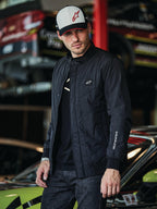 Alpinestars Precedent Jacke, schwarze Steppjacke im Formel-1-Stil, mit einem ikonischen schrägen Steppmuster, Logo-Patch auf der Brust und weißer Ärmelstickerei, getragen mit einer passenden Alpinestars-Kappe in einer Motorsport-Garage.