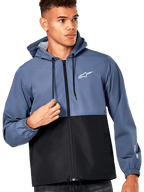 Männliches Model trägt die Alpinestars Speeder Windbreaker-Jacke, Farbblock-Design in Schieferblau und Schwarz, Kapuze mit schwarzen Kordelzügen, weißes Logo auf der Brust, lässiger, vom Motorsport inspirierter Windbreaker