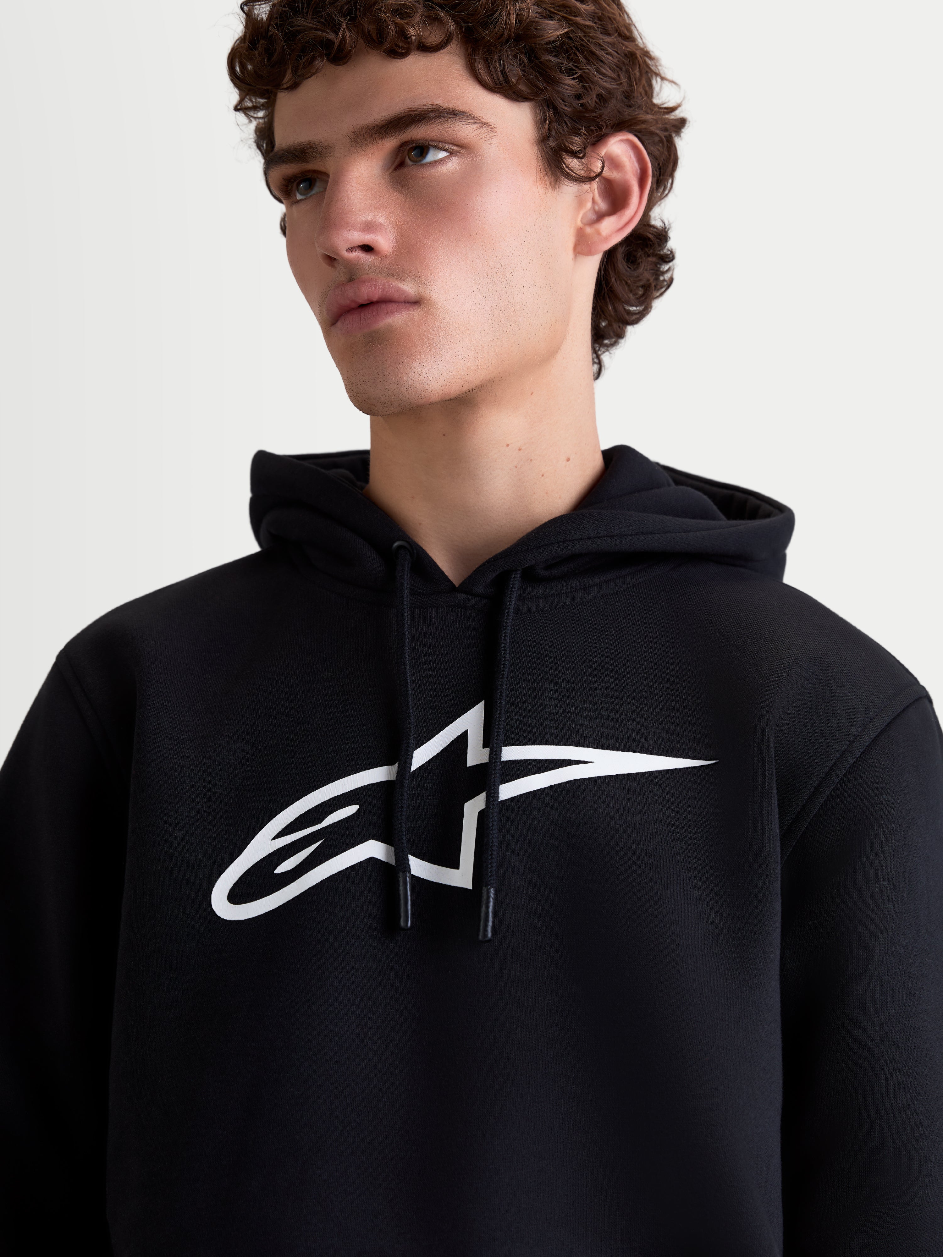 Alpinestars Ageless V3 Hoodie, Herren-Sportswear-Kapuzenpullover aus Fleece, Schwarz und Weiß, Detailansicht am Model mit großem weißem Astars-Logo auf der Brust und Kapuze mit Kordelzug.
