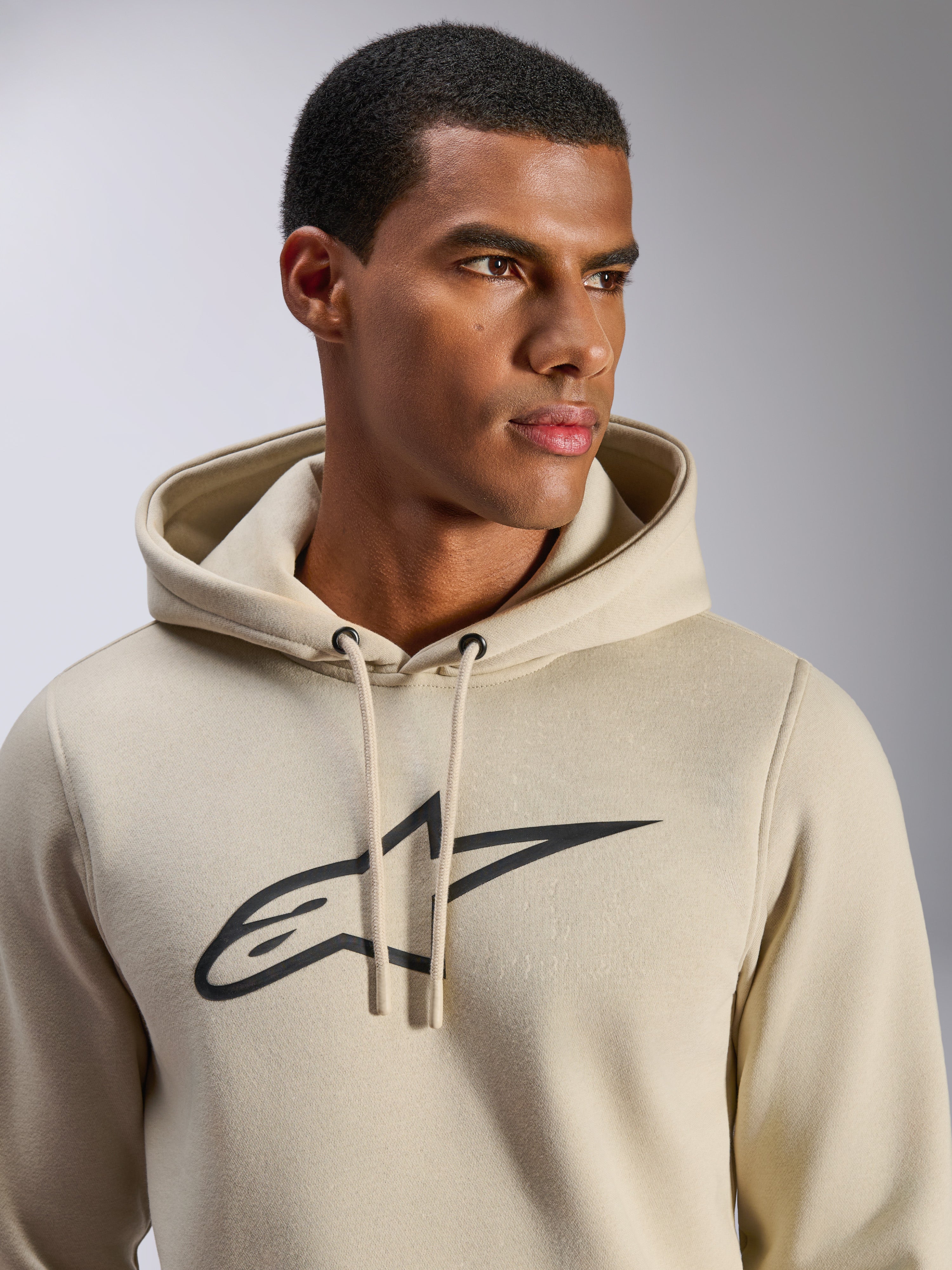 Alpinestars Ageless V3 Hoodie, casual sportswear, off-white with black embroidered Alpinestars star logo, drawstring hood, close-up view on model, soft fleece, modern motorsport-inspired design
