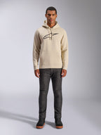 Alpinestars Ageless V3 Hoodie, casual sportswear, Off White with black embroidered logo, worn by a man with dark grey pants and black sneakers, featuring a drawstring hood and kangaroo pocket, modern motorsport-inspired design