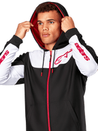 Alpinestars Sessions V3 Hoodie, Schwarz, Weiß und Rot, Kapuzenjacke mit Reißverschluss, getragen von einem Mann mit aufgesetzter Kapuze, rote Alpinestars-Logos auf der Brust und den Ärmeln, lässige Motorsport-Streetwear