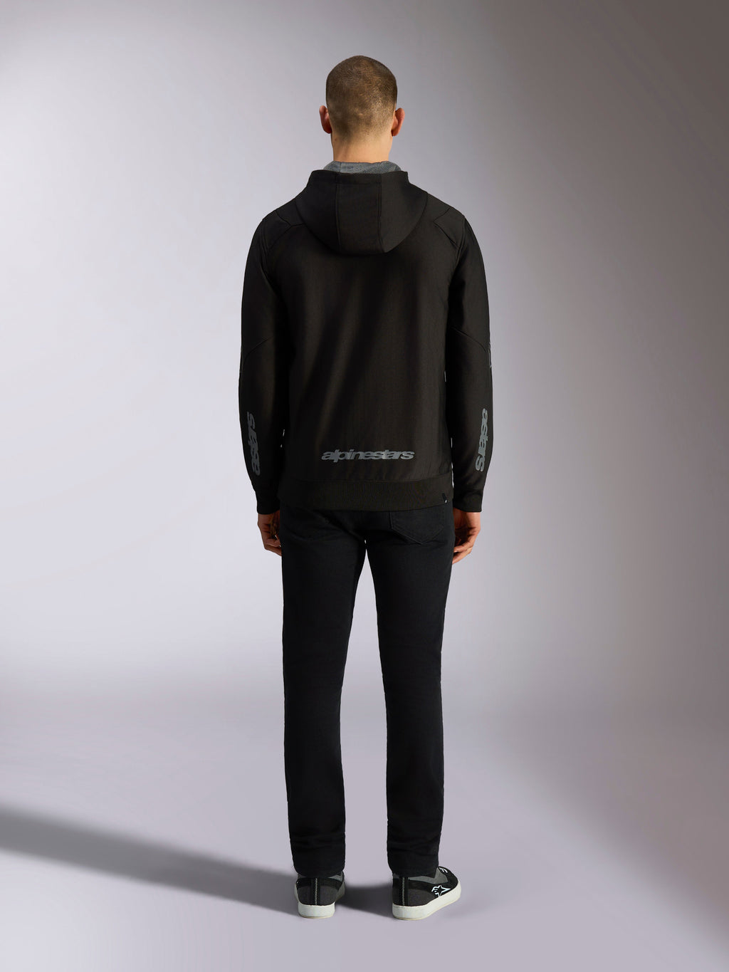 Rückansicht des Alpinestars Sessions V3 Hoodie in Schwarz-Grau, mit einer grauen Alpinestars-Wortmarke am unteren Rücken und Astars-Logos auf den Unterarmen, getragen von einem Model mit schwarzer Hose, lässige vom Motorsport inspirierte Streetwear