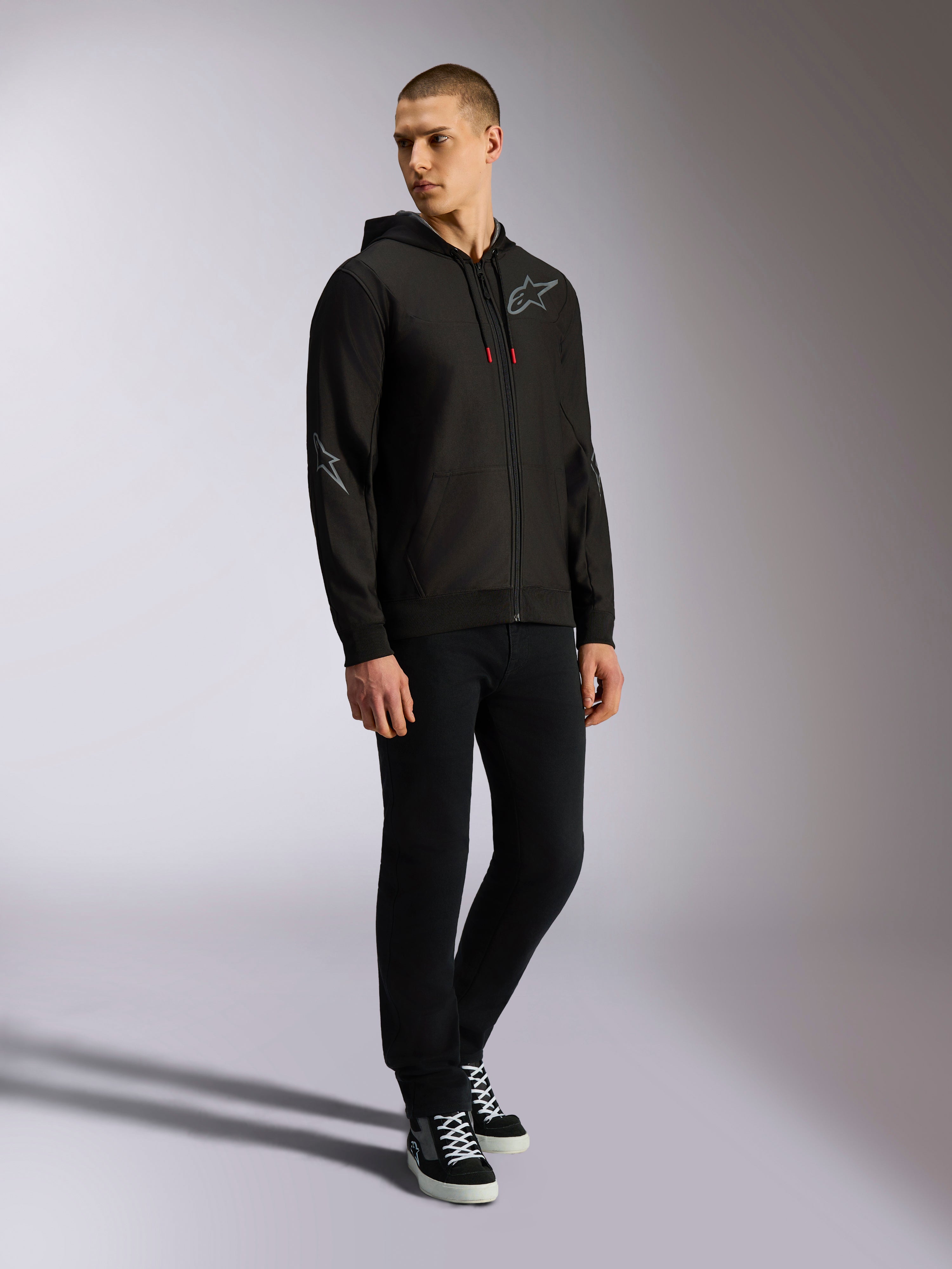Alpinestars Sessions V3 Hoodie, Kapuzenjacke mit Front-Reißverschluss aus Polyfleece, Schwarz und Grau, graue Logos auf Brust und Ärmeln, Kapuze mit Kordelzug und roten Kordelenden, Ganzkörperansicht eines Models in lässiger Motorsport-Streetwear