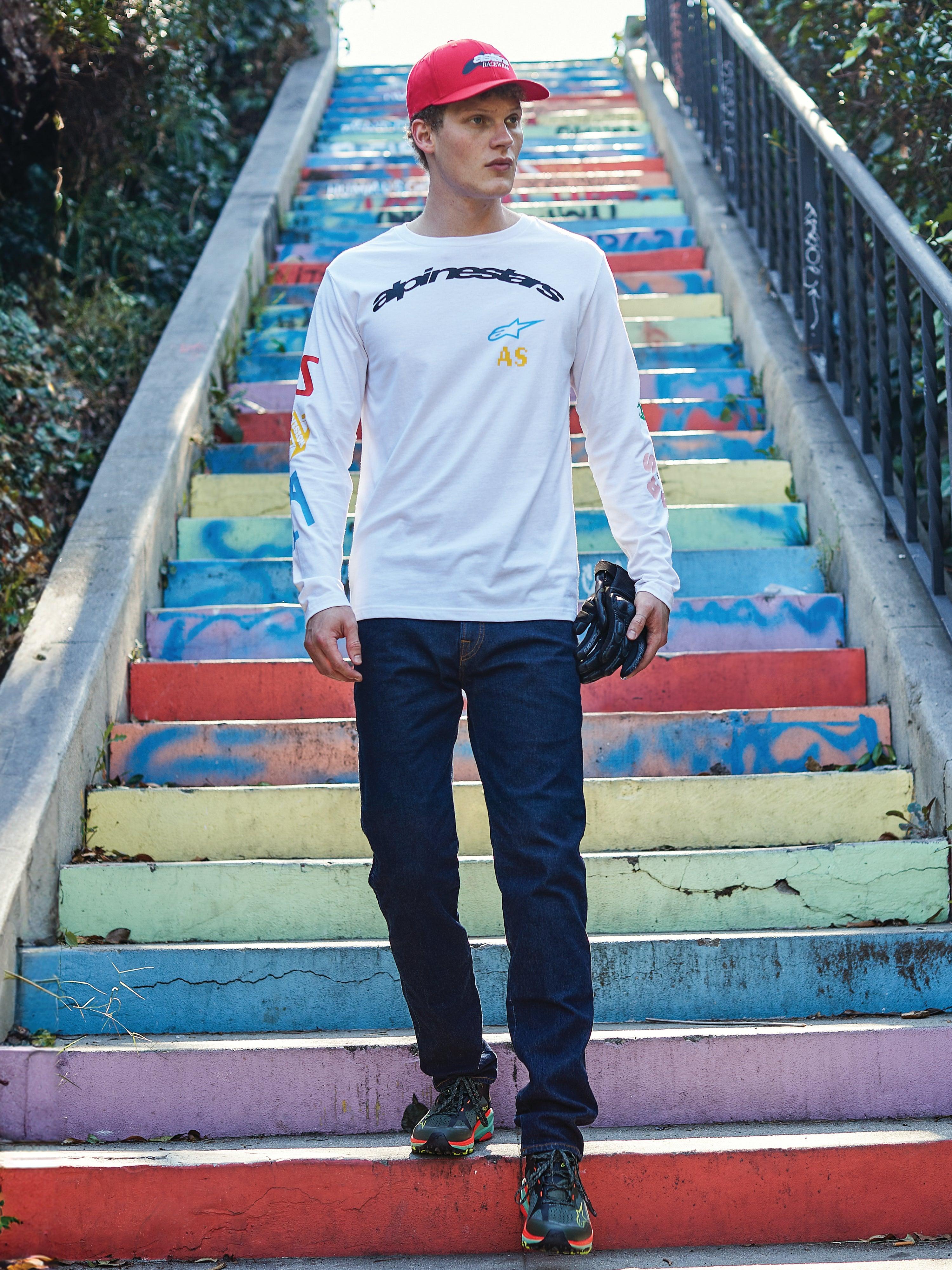 Alpinestars Brundo CSF Langarmshirt, weißes Shirt mit schwarzem Alpinestars-Logo und bunten Grafikdrucken, getragen von einem Model auf einer bunten Treppe, kombiniert mit dunkelblauer Jeans und einer roten Kappe, lässige Motorsport-Streetwear.
