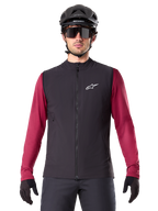 Alpinestars A-Dura Thermoweste, schwarze isolierte Radweste, getragen von einem Modell mit weinrotem Langarmtrikot, schwarzem MTB-Helm und Sonnenbrille, mit weißem Logo auf der Brust, leichter Wärmeschutz fürs Mountainbiken