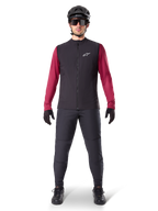 Alpinestars A-Dura Thermoweste, schwarze Radweste getragen über einem weinroten Langarmtrikot, kombiniert mit dunkelgrauer MTB-Hose, schwarzem Helm und Sonnenbrille, technische Mountainbike-Bekleidung für Fahrten bei kaltem Wetter