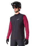 Alpinestars A-Dura Thermoweste, Schwarz, Radsport-Gilet, getragen über einem weinroten Langarmshirt, mit weißem Logo auf der Brust, kombiniert mit schwarzem MTB-Helm und Sonnenbrille, isolierende Performance-Schicht für das Mountainbiken