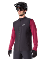 Alpinestars A-Dura Thermoweste, Schwarz, Radsport-Gilet, getragen über einem weinroten Langarmshirt, mit weißem Logo auf der Brust, kombiniert mit schwarzem MTB-Helm und Sonnenbrille, isolierende Performance-Schicht für das Mountainbiken