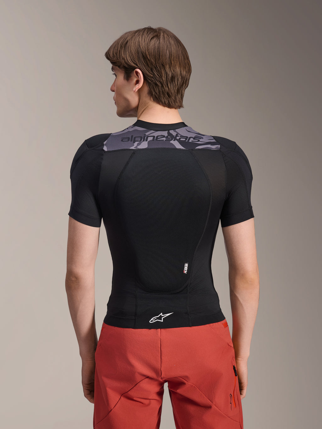 Alpinestars A-Motion Plasma Protection Jacket - Short Sleeve, Black with Grey Camo accents on the upper back, back view showing integrated back protector and white Astars logo at the hem, worn with red shorts, technical mountain bike protection gear