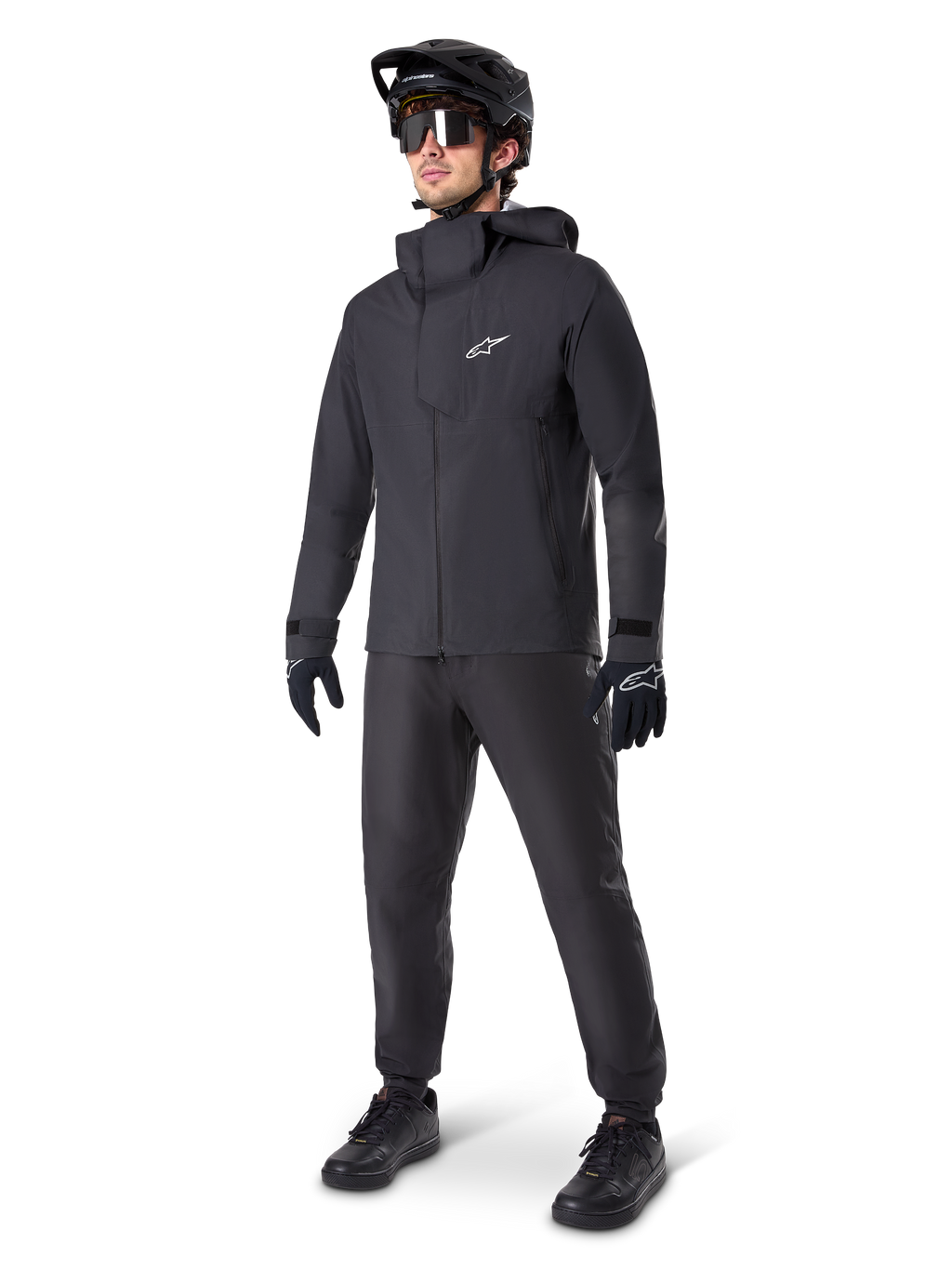 Männlicher Mountainbiker in schwarzen Alpinestars A-Dura Waterproof Pants, kombiniert mit einer passenden schwarzen wasserdichten Kapuzenjacke, schwarzem MTB-Helm mit Visier, dunkler Radsportbrille und schwarzen Handschuhen, komplette Profi-Ausrüstung für Trail-Fahrten bei jedem Wetter